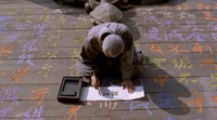 A man kneels as he writes black calligraphy on white paper, with an inkstone holding the paper down, on top of painted calligraphic text carved into a wood floor.