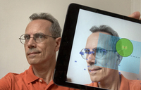 Color photo, a man with glasses is holding an iPad at arms-­length away from their face. His face is also shown on the AUMI screen.