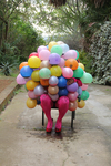 A figure engulfed in multicolored balloons and wearing hot pink stockings sits in a chair. The balloons have leaked liquid onto the ground at its feet