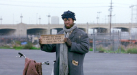 A tramp at an airport holding a sign written on a piece of cardboard