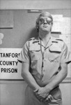 A young blond man in a militaristic “guard” uniform and mirror shades, leaning against a wall. The words “Stanford County Prison” are visible on a sign near his elbow.