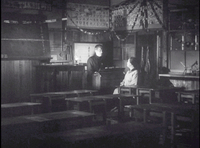 Empty desks create the foreground. Two people are in the corner of an empty room with one light shining towards them and paper chains draping above them. One person stands behind a podium and the other sits directly in front of the podium. Calligraphy hangs on the wall above them.