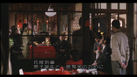 Image of a restaurant with several people standing and seated. There is calligraphy in a subtitle as well as on the back wall, on red and pink banners.