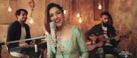 Medium shot of Neeti Mohan singing a version of her song “Nainowale Ne,” from Padmaavat, on a music video for the album T-­Series Acoustics. Standing at a mic, she smiles at the camera; in the background, on either side of her, are a keyboardist and a guitarist.