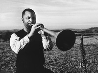 Blackand-white image depicting a man sitting in an open field, playing a long straight wind instrument intensely with his eyes closed, its flared bell to the viewer’s right.