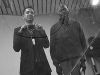 Muhal Richard Abrams and Vijay Iyer standing beside one another, as Iyer takes their photograph reflected through a dusty mirror.