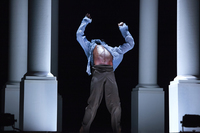 A male figure is framed by four white columns. He arches back in a fast, dramatic throw of his body, his head barely visible as his blue shirt flies up and reveals his torso. He plants his feet as his arms stretch overhead.