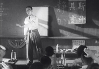 A teacher addresses the classroom in front of a whiteboard set against the chalkboard with white calligraphy and paper attached to it with white calligraphy, in black and white cinematography.