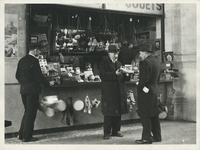 Georges demonstrates a toy car to a gentleman at the Confiserie et Jouets toy kiosk while another man browses.