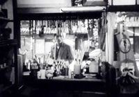 A man look towards the viewer across a counter full of calligraphy supplies, in black and white cinematography